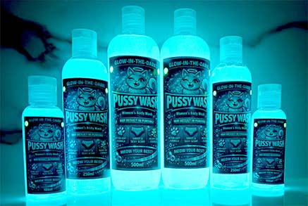 Six glow-in-the-dark bottles of Pussy Wash personal care products arranged in a row against a dark, black background with a faint silhouette.