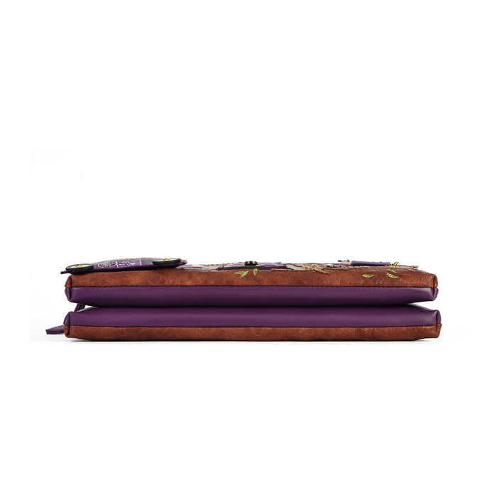 A closed zip-around wallet or planner shown from the side on a plain white background. The exterior is a textured brown faux-leather with a floral vine pattern in purple, green, and gold, and the interior lining is a solid deep purple.