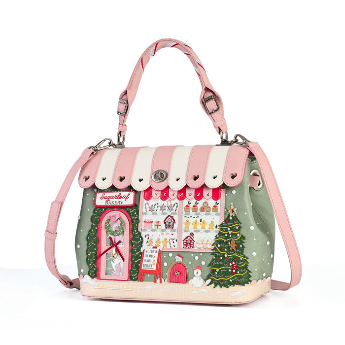 A pink and sage green handbag designed to look like a whimsical Christmas village shop, featuring a pink and white striped awning, a decorated Christmas tree, a red Santa's sleigh sign, and a snowy base, with a pink shoulder strap and top handle.