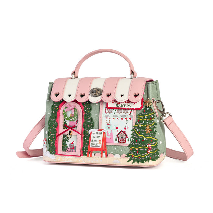 A pink and green Christmas-themed handbag with a top handle, shoulder strap, and festive printed details including a bakery sign, Christmas tree, and window illustration, shown on a plain white background.