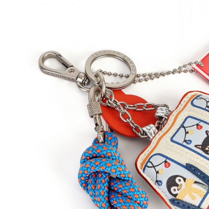 A close-up of a keychain set on a white background, featuring a silver-tone metal clasp and ball chain, a red heart-shaped charm, a blue and orange knotted cord tassel, and a small fabric pouch with cartoon penguin artwork.
