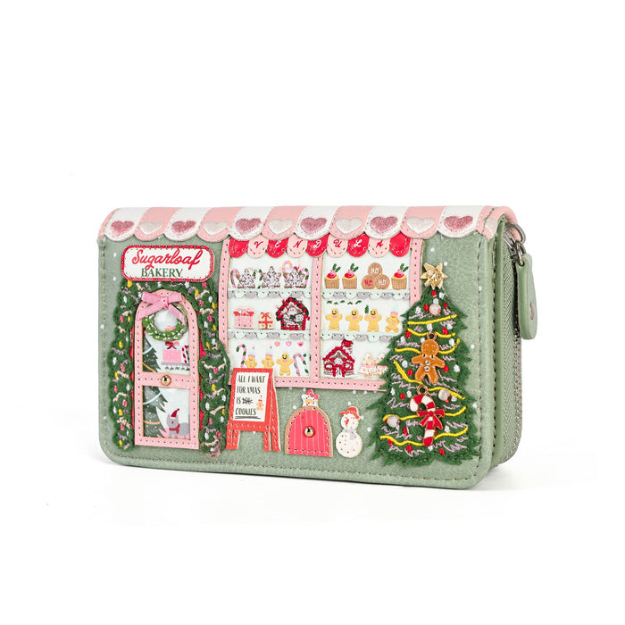 A light sage green zip-around clutch wallet decorated with a detailed Christmas bakery scene. The design features pink and white striped awning, a green wreath door, a bakery window display with pastries, a gingerbread man, a snowman, and a decorated Christmas tree.