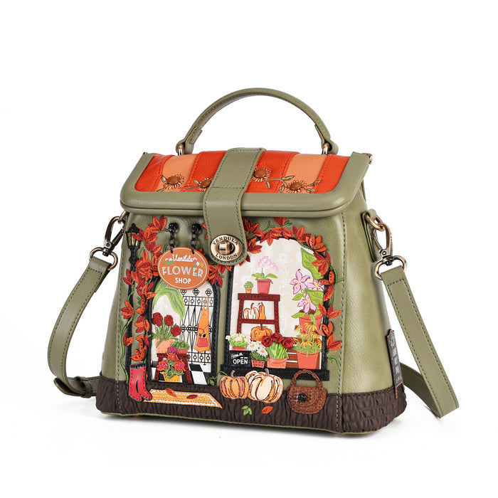 An olive green handbag with a top handle and shoulder strap, featuring a detailed autumn-themed patchwork design on the front flap with a flower shop illustration, pumpkins, and orange leafy borders.