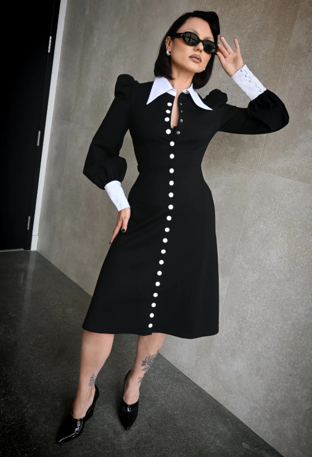 Woman wearing a black dress with white buttons against a gray wall.
