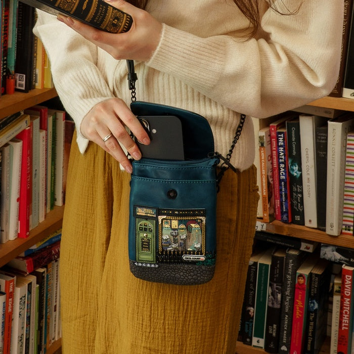 Person holding a blue crossbody bag with a phone case in front of a bookshelf.