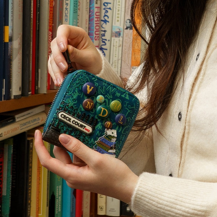 Person holding a blue wallet with decorative elements in front of a bookshelf