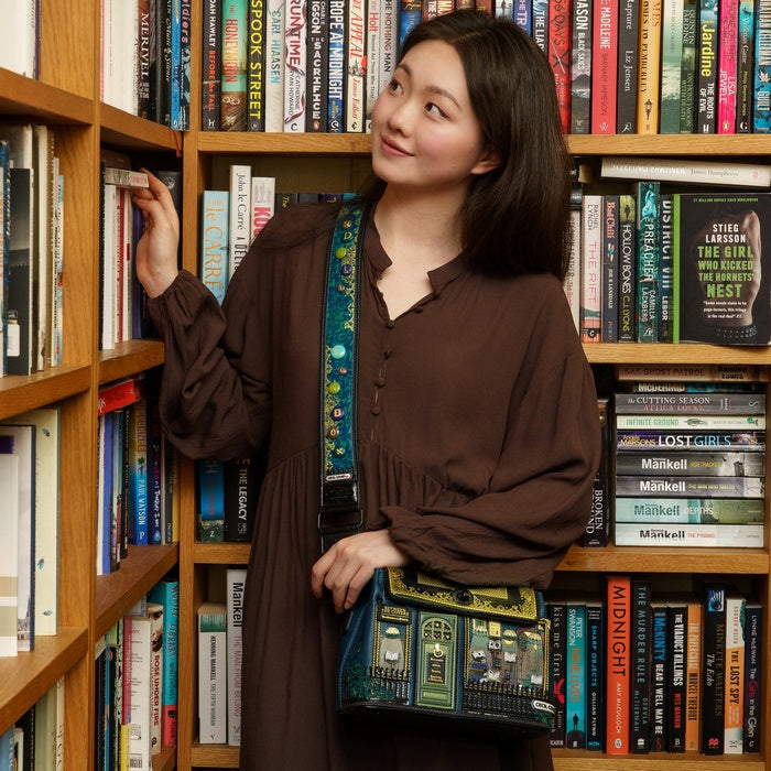 Woman in a brown dress holding a book-themed handbag in front of a bookshelf.