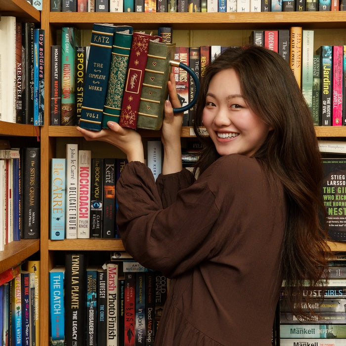 Woman holding a book-shaped mug in front of a bookshelf filled with books.
