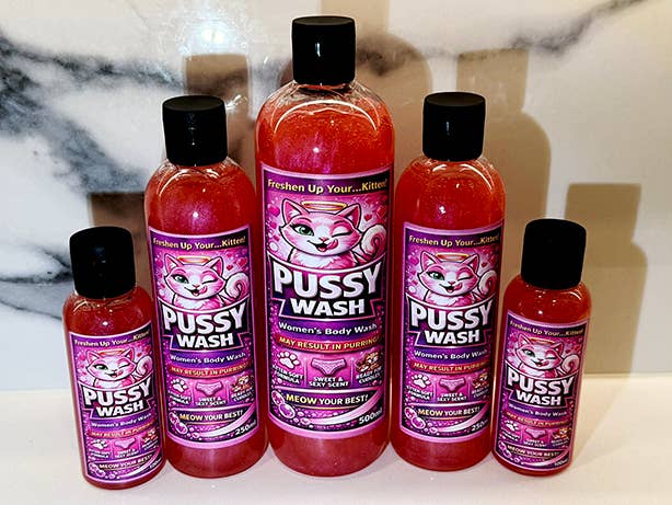 Five bottles of Pussy Wash women's body wash in varying sizes, arranged in a pyramid on a white marble surface. The bottles are filled with a pinkish-red liquid and feature black caps and pink and purple labels with a cartoon cat illustration.