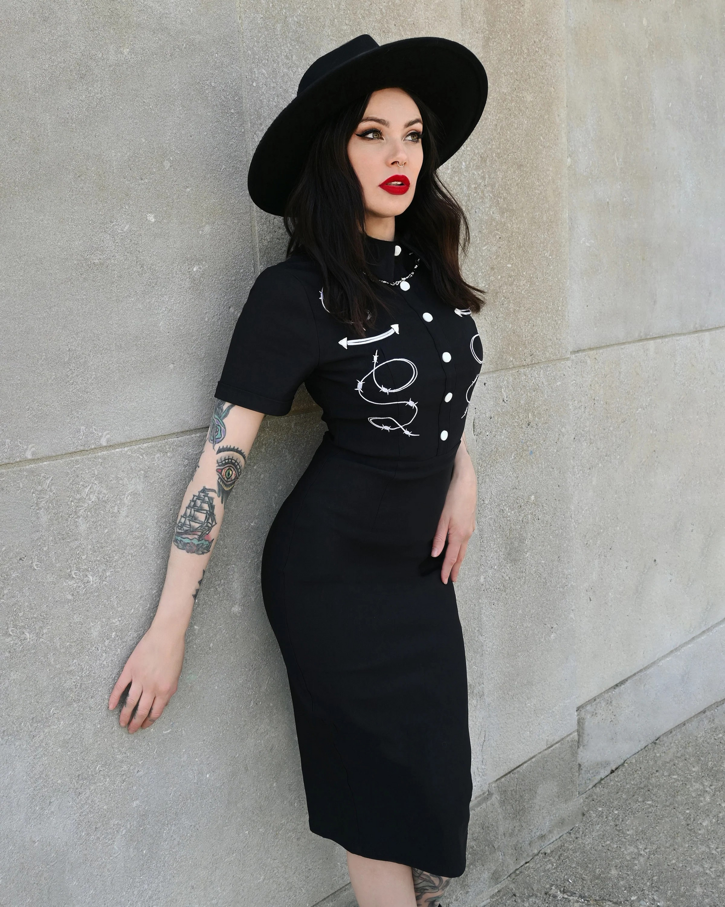 Woman wearing a black outfit with white polka dots and a black hat against a gray wall.
