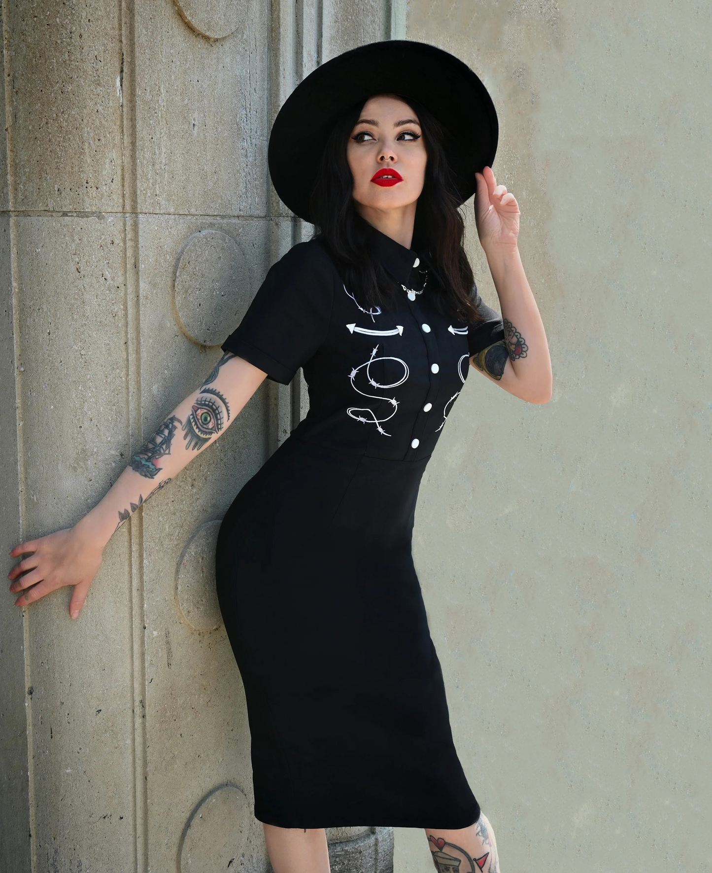 Woman wearing a black dress with white designs and a black hat against a concrete wall.