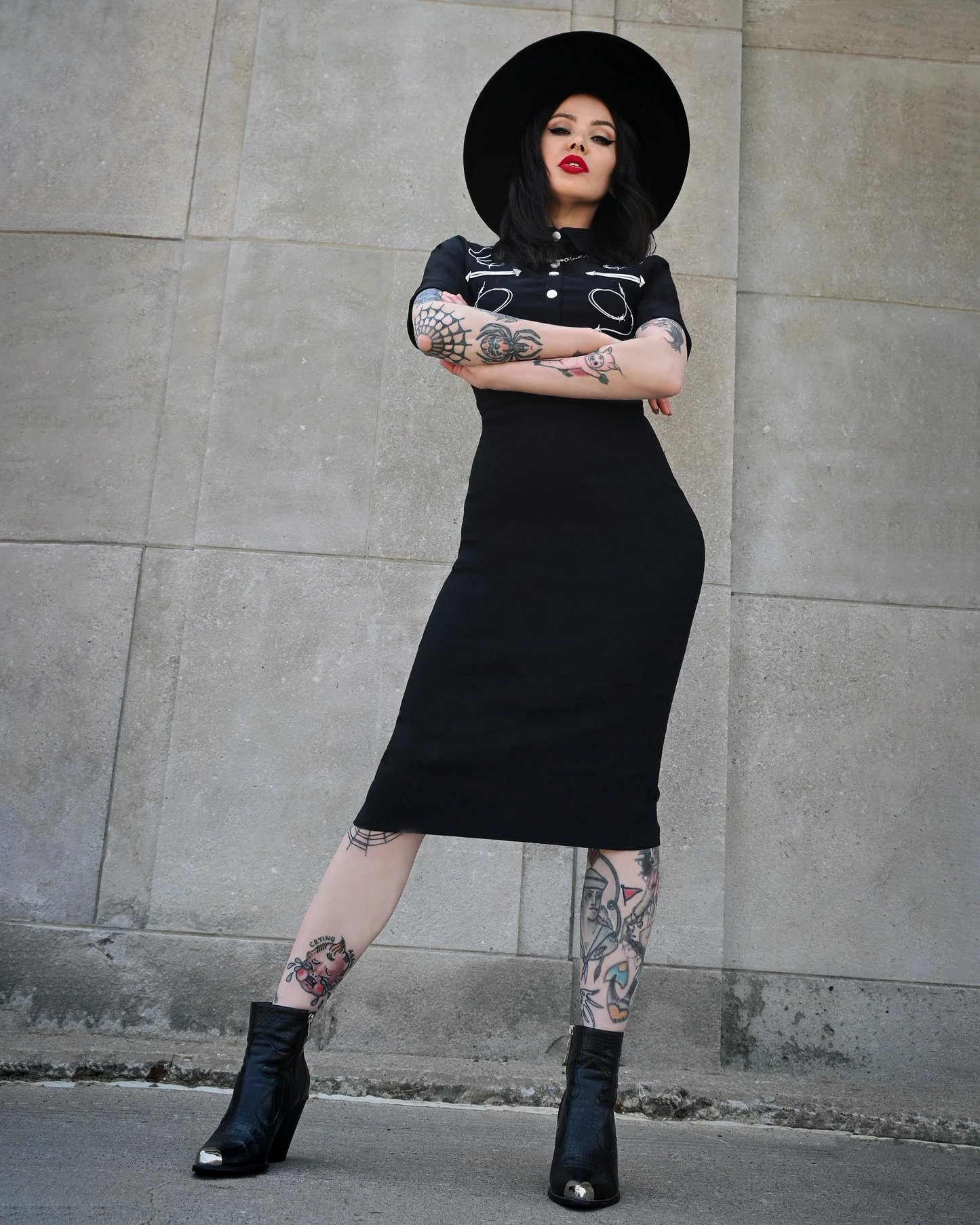 Woman in a black dress and hat with tattoos standing against a gray wall.