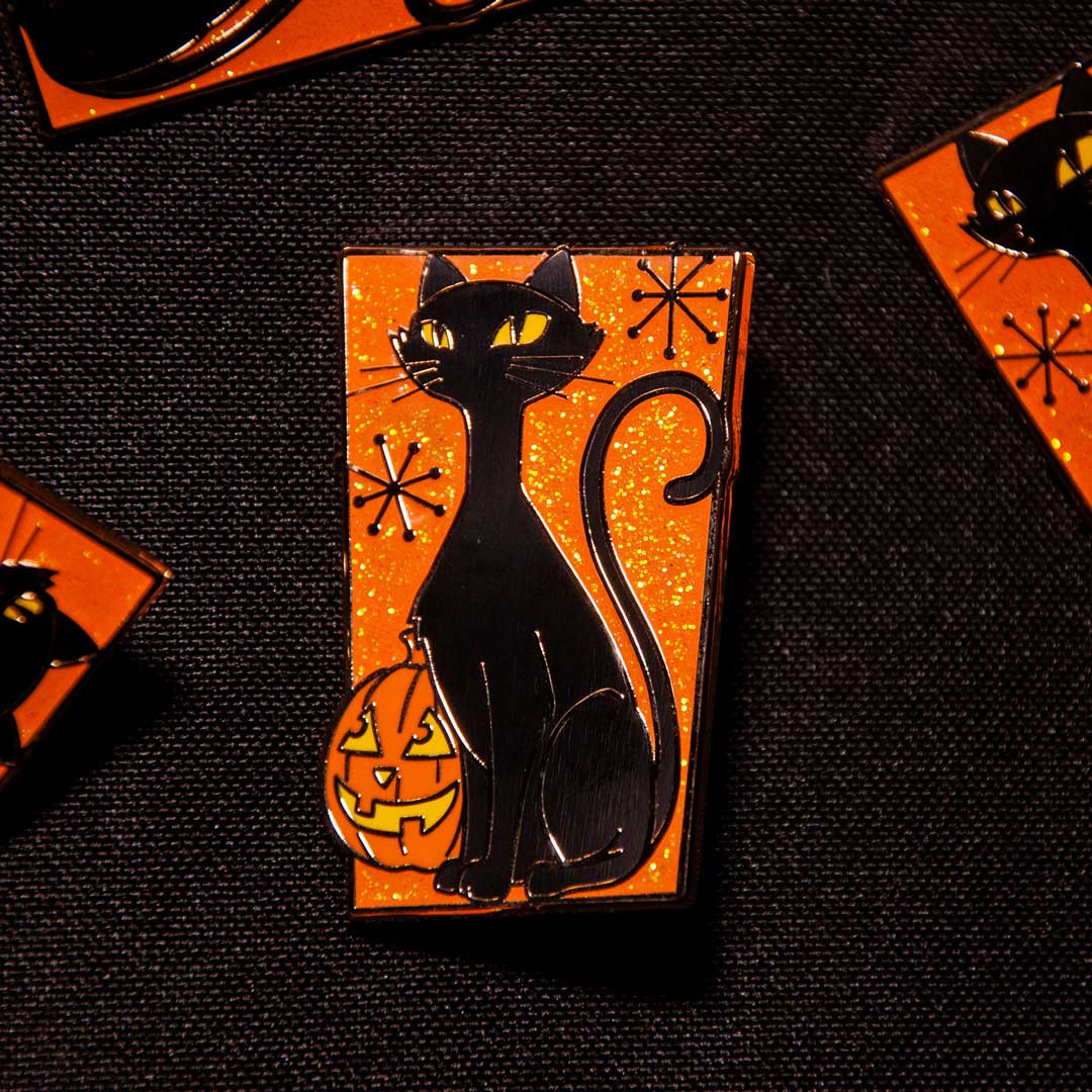 A rectangular enamel pin featuring a black cat sitting on an orange glittery background with a carved jack-o'-lantern and small black starburst motifs, displayed on a dark textured surface.