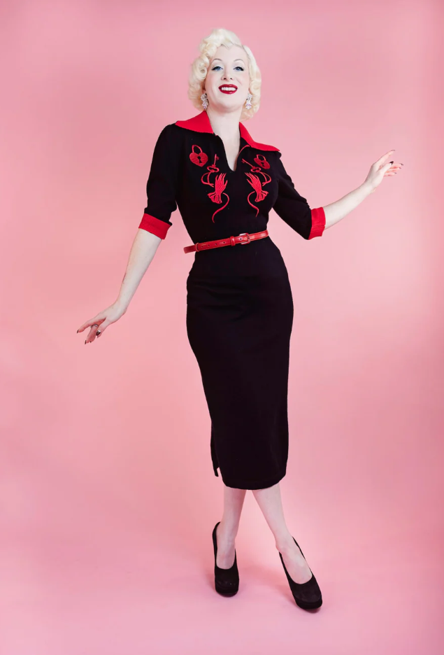 Woman in a black dress with red accents posing against a pink background
