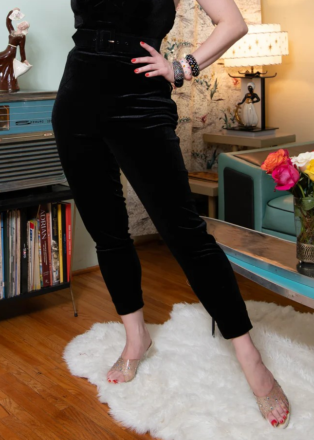 Person wearing black pants standing on a white rug in a room with books and decorative items.