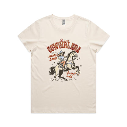 A white graphic t-shirt featuring a retro western illustration of a cowgirl riding a horse. The text reads 'In my COWGIRL ERA' in arched red letters, 'Always Sassy' in red script, and 'Always Wild' in red script, with the cowgirl wearing a cowboy hat and boots while riding a rearing horse.
