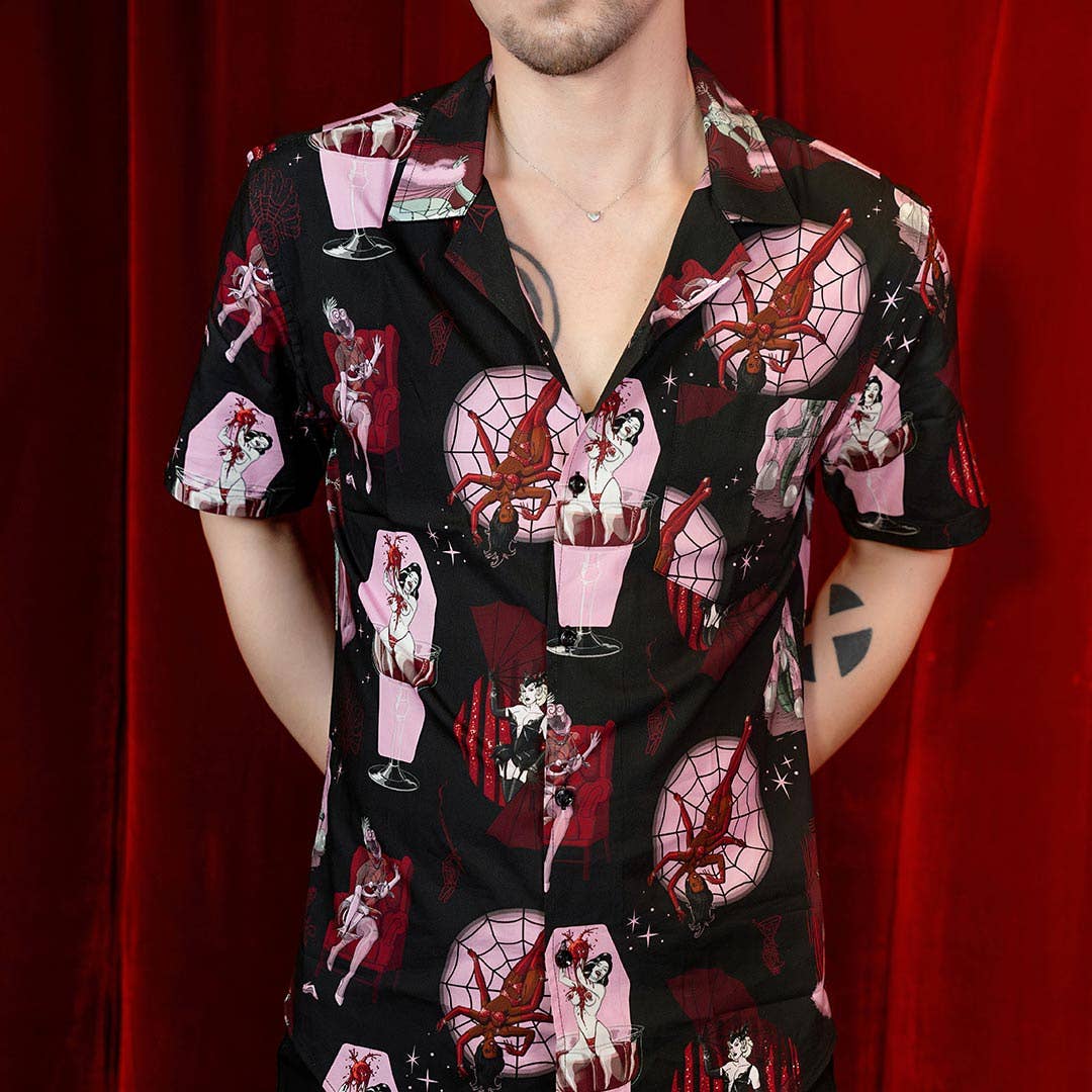 A man wearing a black short-sleeve button-up shirt with a pink and red gothic anime print, posed against a red curtain background.