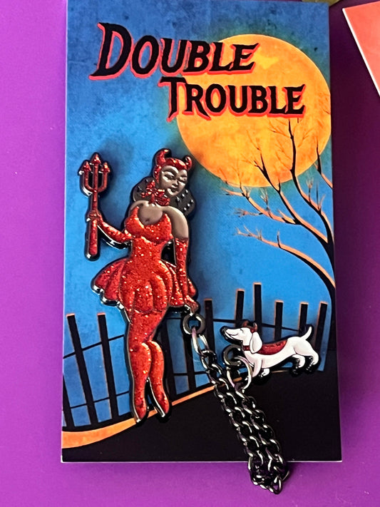 A collectible enamel pin featuring a red devil character and a white dog on a black chain, mounted on its original illustrated backing card titled 'Double Trouble' against a purple background.