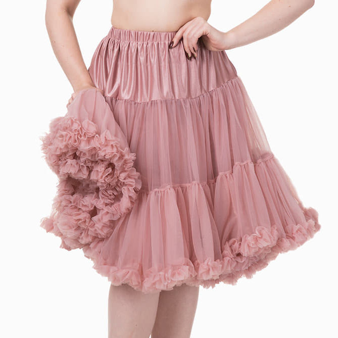 A woman modeling a pink tiered tulle skirt with a ruffled pom-pom accessory, paired with a black bralette and black heels, against a plain white background.
