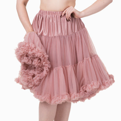 A woman modeling a pink tiered tulle skirt with a ruffled pom-pom accessory, paired with a black bralette and black heels, against a plain white background.