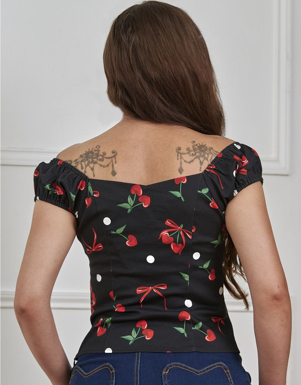 Woman wearing a black top with red cherries and green leaves, standing against a white wall.