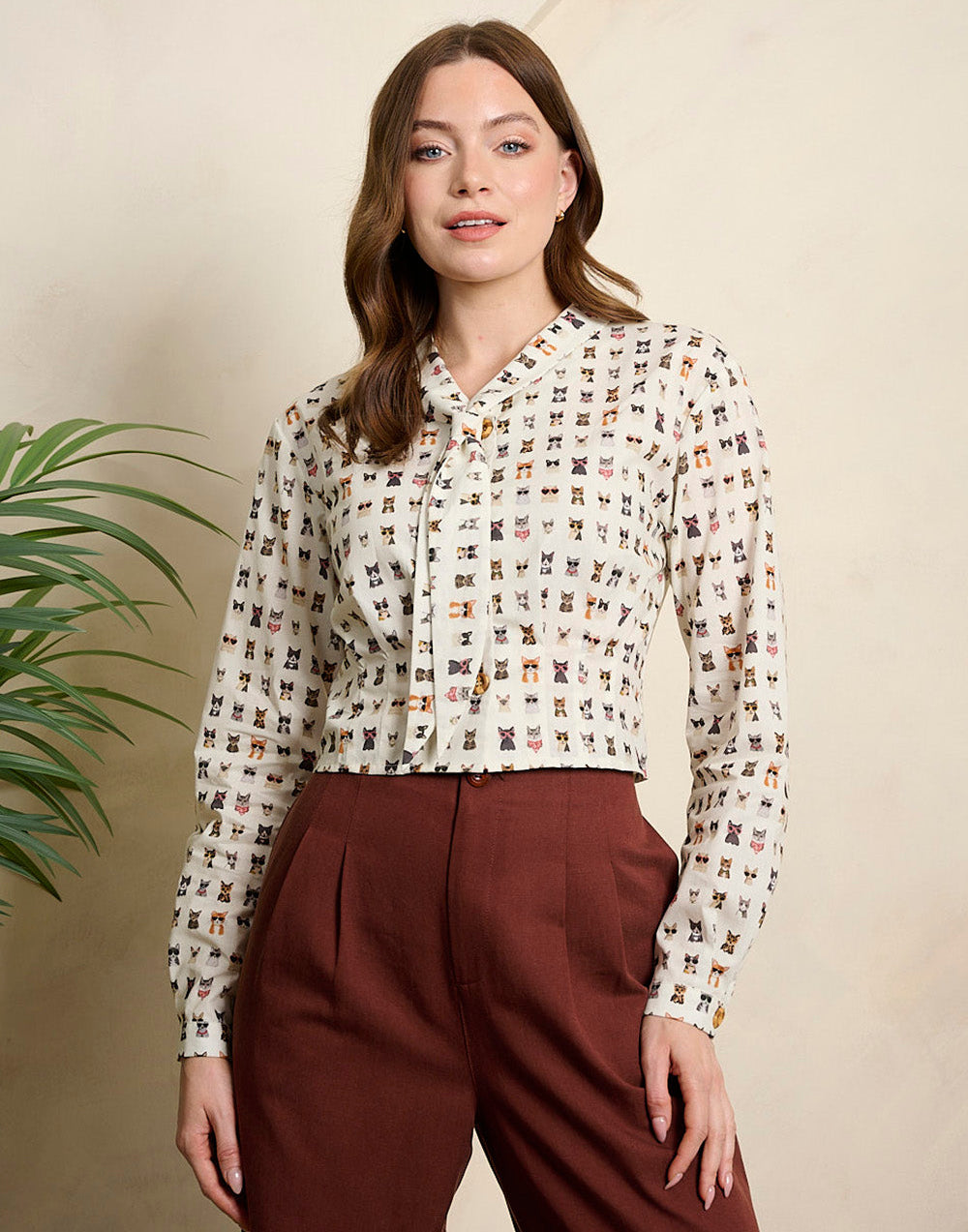 A woman modeling a long-sleeve button-up blouse with a small repeating multicolor print on a light beige background, paired with dark brown trousers, set against a warm beige wall with a green plant leaf visible on the left.