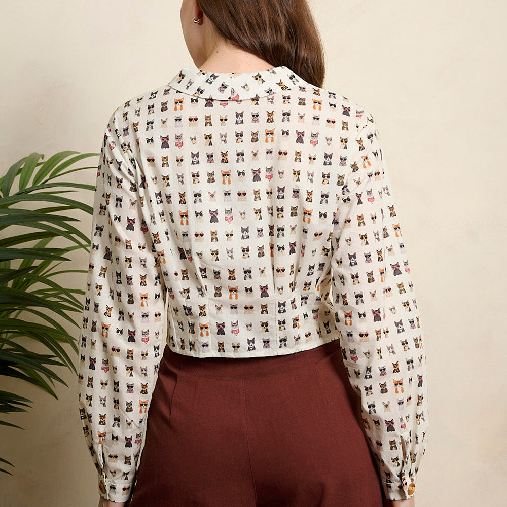 A woman modeling a long-sleeve button-up blouse with a small repeating multicolor print on a light beige background, paired with dark brown trousers, set against a warm beige wall with a green plant leaf visible on the left.