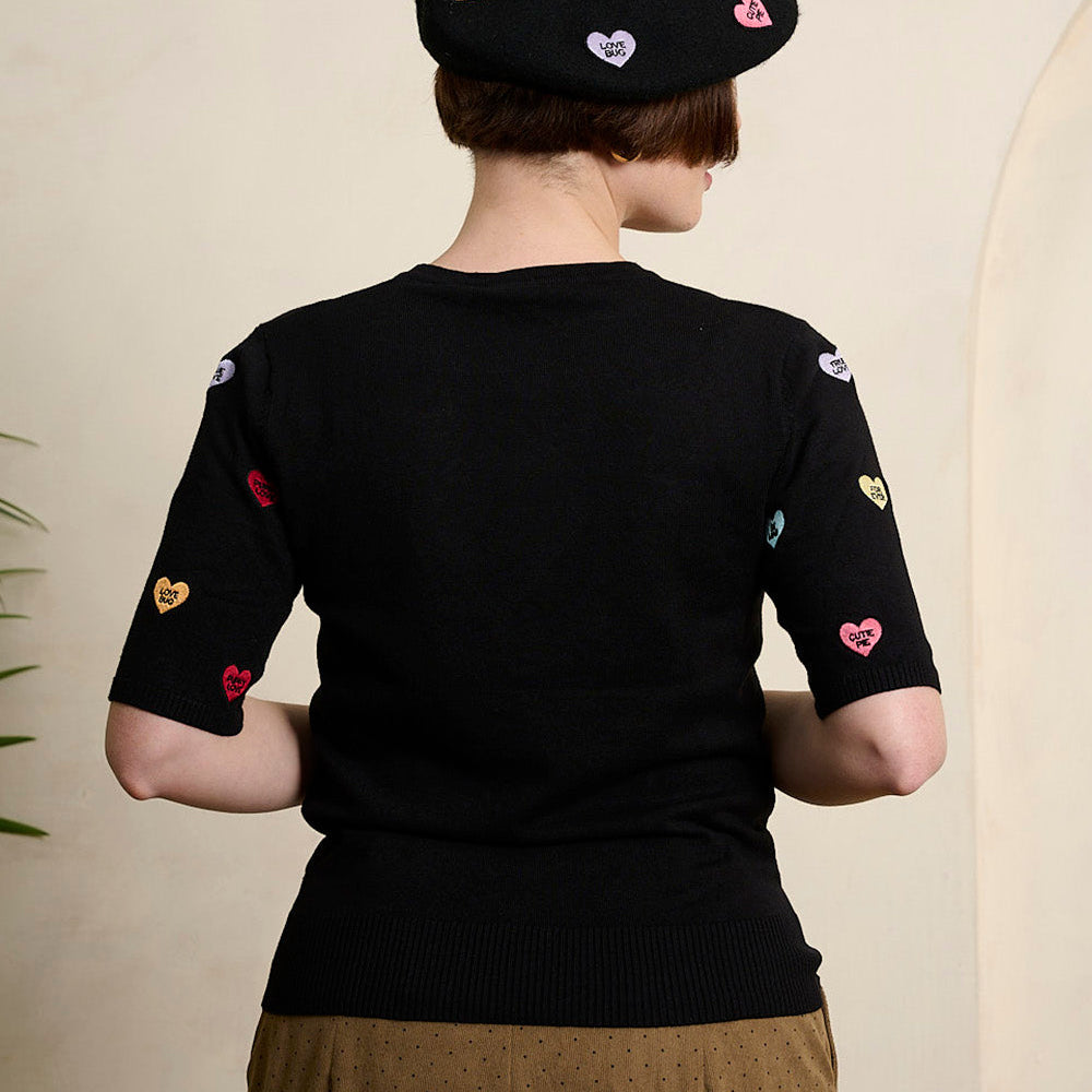 A person shown from the back wearing a black short-sleeve knit top with colorful embroidered heart motifs on the sleeves, a black beret with heart patches, and brown wide-leg trousers, standing indoors against a light beige wall.