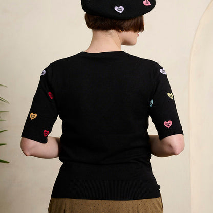 A person shown from the back wearing a black short-sleeve knit top with colorful embroidered heart motifs on the sleeves, a black beret with heart patches, and brown wide-leg trousers, standing indoors against a light beige wall.