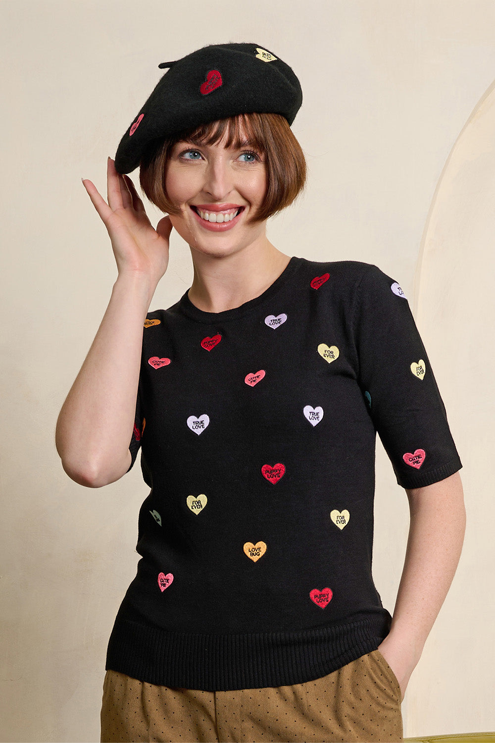 A smiling woman with a short brown bob haircut wears a black knit beret and a black short-sleeve sweater decorated with small colorful heart patches in red, gold, and white. She is wearing brown corduroy trousers against a plain beige studio background.