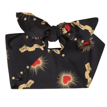 A black satin bow headband featuring a repeating pattern of gold-toned classical statues, red hearts with gold rays, and small gold stars, shown on a plain white background.