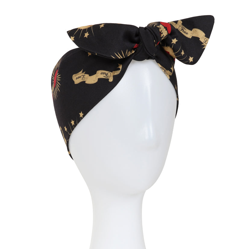 A black silk-like headscarf with a bow, featuring a gold horse and rider pattern and small red starburst accents, displayed on a white mannequin head against a white background.