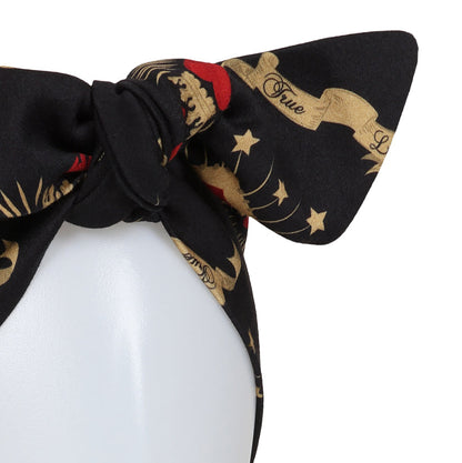 A black silk scarf with a gold and red baroque-style print, tied in a large bow on a white mannequin head against a white background.