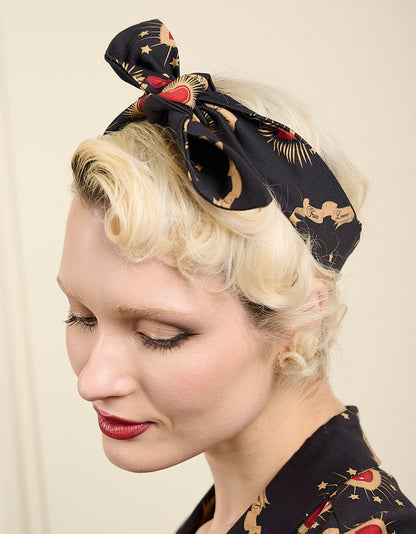 A woman with vintage-style blonde hair wearing a black silk headscarf with a bow, decorated with gold stars and red hearts, paired with a matching black blouse.