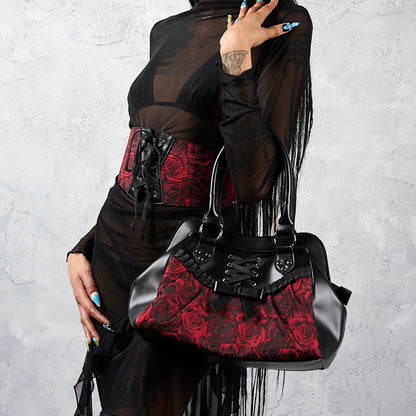 A model wearing a black sheer long-sleeve top and skirt with a red and black lace-up corset, holding a black and red rose-patterned shoulder bag against a textured gray wall.