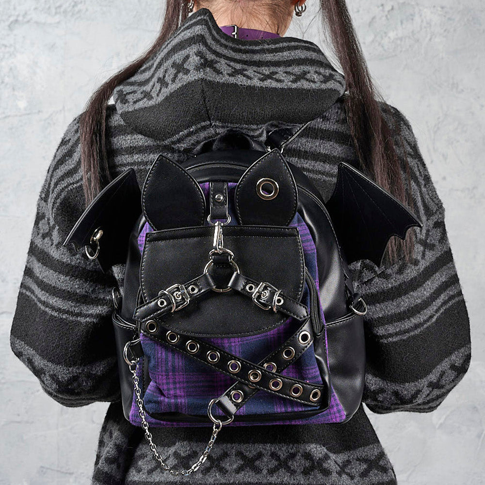 A person viewed from behind wearing a black and purple gothic backpack with bat wings and chain details, paired with a gray patterned sweater against a textured gray wall.