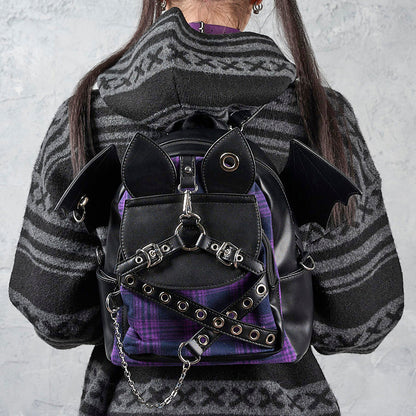 A person viewed from behind wearing a black and purple gothic backpack with bat wings and chain details, paired with a gray patterned sweater against a textured gray wall.
