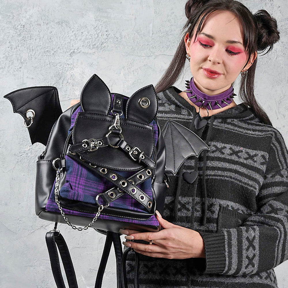 A goth-style model wearing a black and gray patterned sweater and purple choker holds a black and purple gothic backpack with bat wings and metal chain details against a textured gray wall.