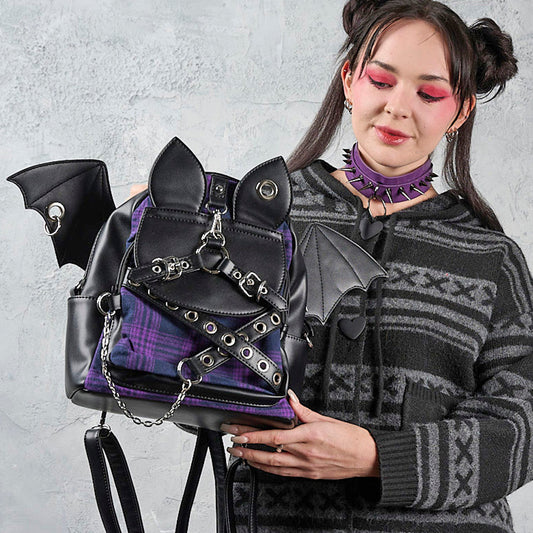 A goth-style model wearing a black and gray patterned sweater and purple choker holds a black and purple gothic backpack with bat wings and metal chain details against a textured gray wall.