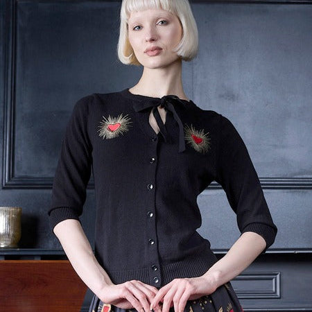 A woman with a short blonde bob stands indoors wearing a black button-front cardigan with embroidered heart motifs and a dark patterned skirt. The background is a dark paneled wall with a wooden cabinet visible at the lower left.