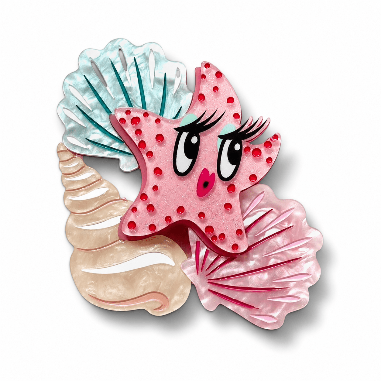 A whimsical starfish-shaped hair clip or brooch featuring a pink starfish with red polka dots, large cartoon eyes, eyelashes, and a small pink nose, layered over three seashell shapes in light blue, beige, and darker pink on a white background.