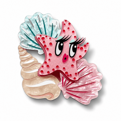 A whimsical starfish-shaped hair clip or brooch featuring a pink starfish with red polka dots, large cartoon eyes, eyelashes, and a small pink nose, layered over three seashell shapes in light blue, beige, and darker pink on a white background.