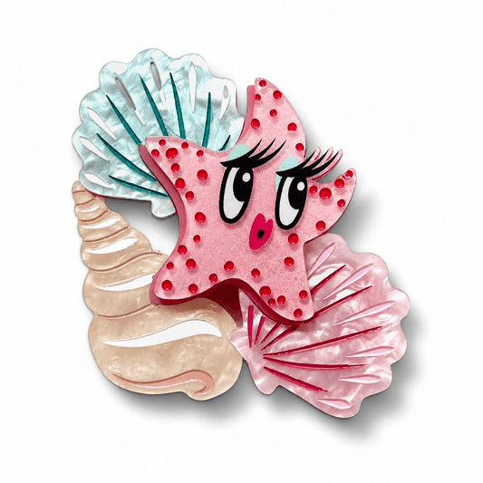 A whimsical starfish-shaped hair clip or brooch featuring a pink starfish with red polka dots, large cartoon eyes, eyelashes, and a small pink nose, layered over three seashell shapes in light blue, beige, and darker pink on a white background.