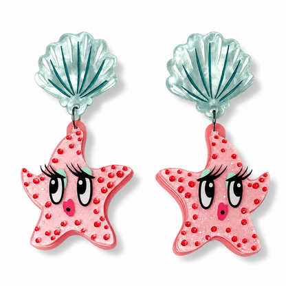 A pair of dangle earrings featuring pink starfish charms with cartoon eyes and red rhinestone accents, hanging from translucent light blue seashell studs on a white background.