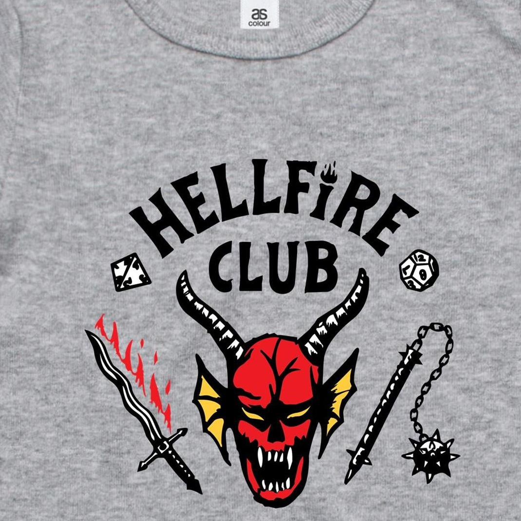 A close-up of a heather gray t-shirt featuring a graphic print with the text 'HELLFIRE CLUB' in black. Below the text is a red horned devil face with yellow wings, flanked by a black-and-red sword on the left and a black spiked chain weapon on the right.