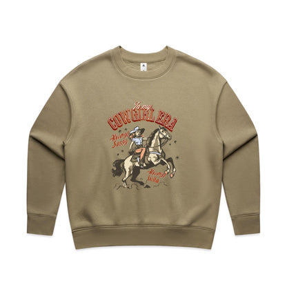A close-up of a cream-colored t-shirt featuring a vintage-style western graphic. The graphic shows a cowboy riding a rearing horse, with retro text reading 'In my COWGIRL ERA', 'Always Sassy', and 'Always Wild' in reddish-brown and black.