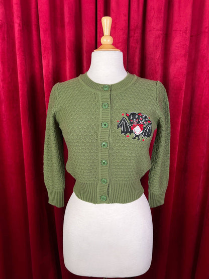 Green cardigan with a decorative patch on a mannequin against a red curtain background