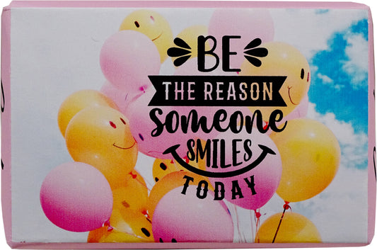 A rectangular product package or box featuring a printed design of pastel pink, yellow, and white balloons with smiley faces against a blue sky. The center has black decorative text reading 'BE THE REASON someone SMILES TODAY'.