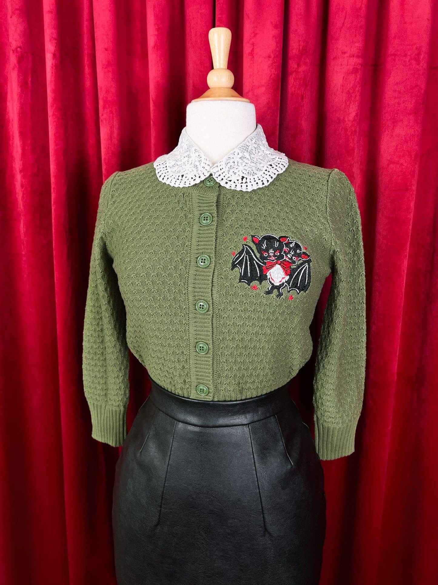 Green cardigan with a decorative emblem on a mannequin against a red curtain background
