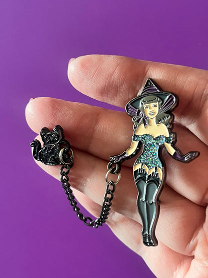 A hand holding a black witch-themed enamel pin set against a solid purple background. The set includes a witch figure with a black hat and dress with teal and gold details, and a small black cat charm connected by a short chain.
